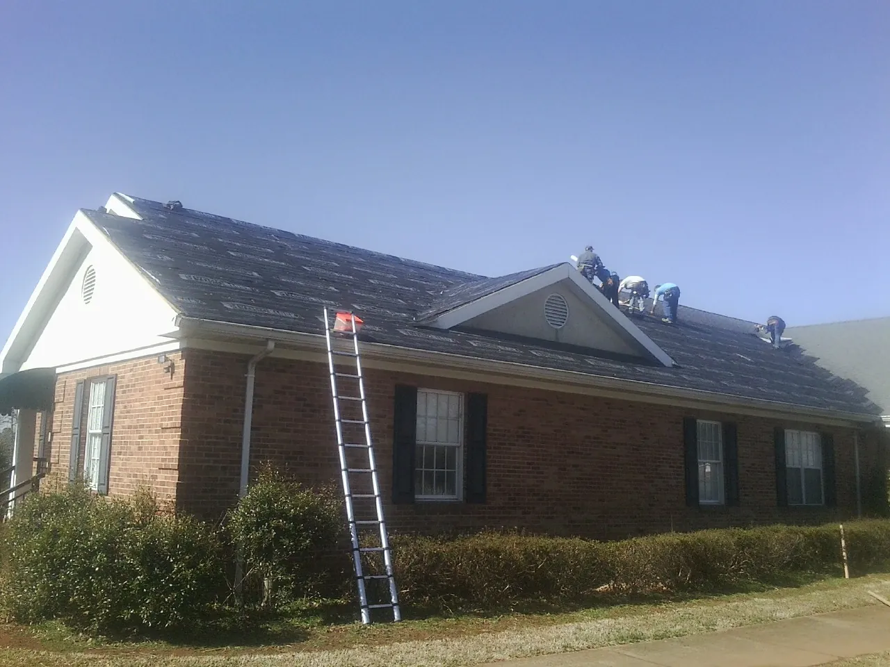 Crew tearing shingles off a brick ranch roof with a ladder set — Metal Roof Repair work by our crew in Dyer
