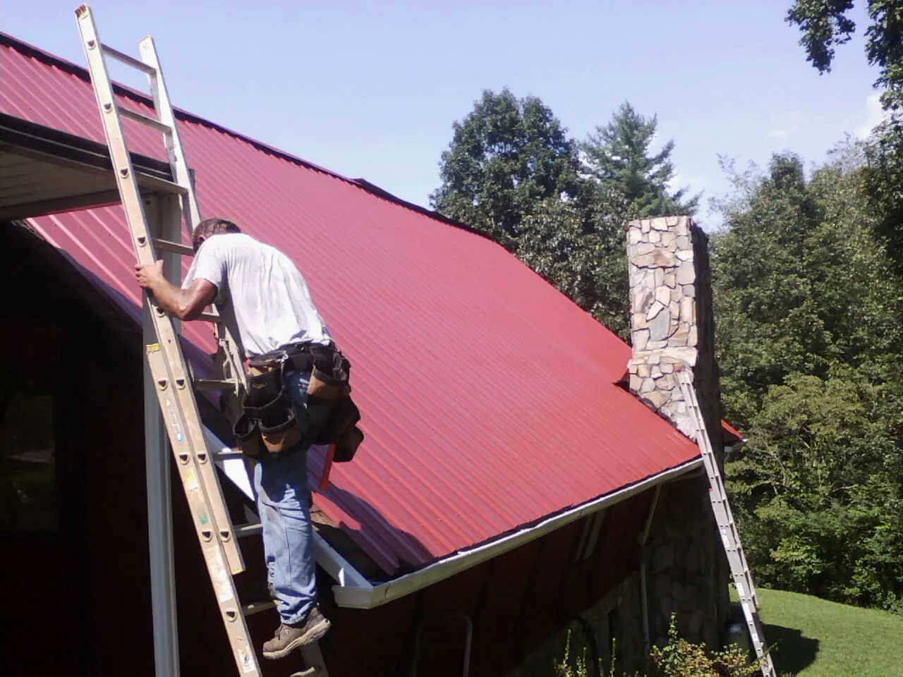 Roofer on a ladder beside a red metal roof and stone chimney — Soffit & Fascia Repair by Roofing Brothers in Dyer